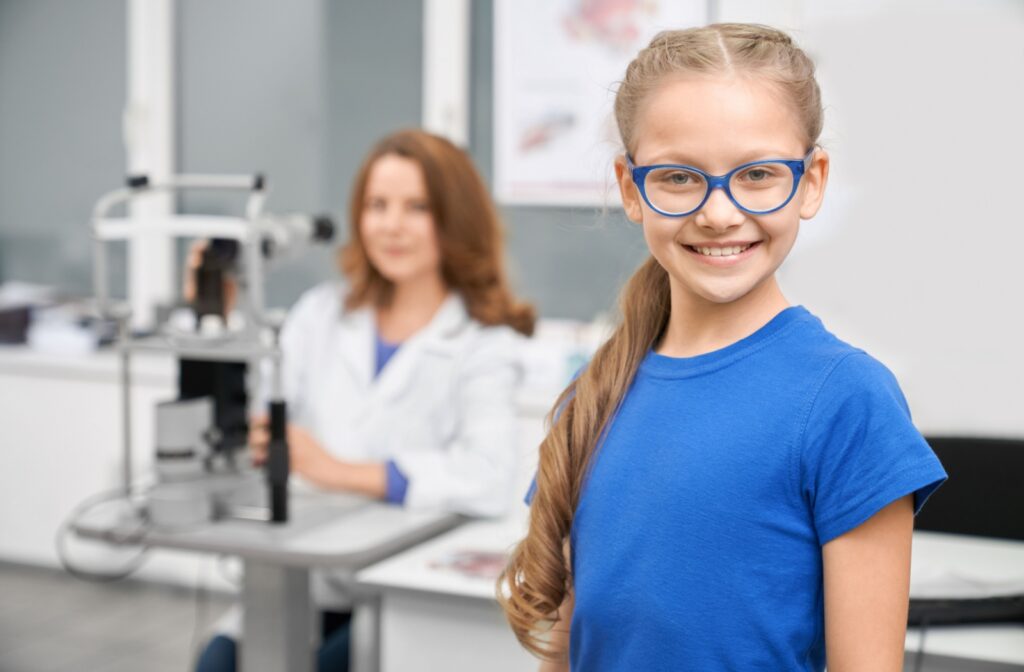 A child with glasses smiling and looking directly at the camera, while an optometrist working on the background.
