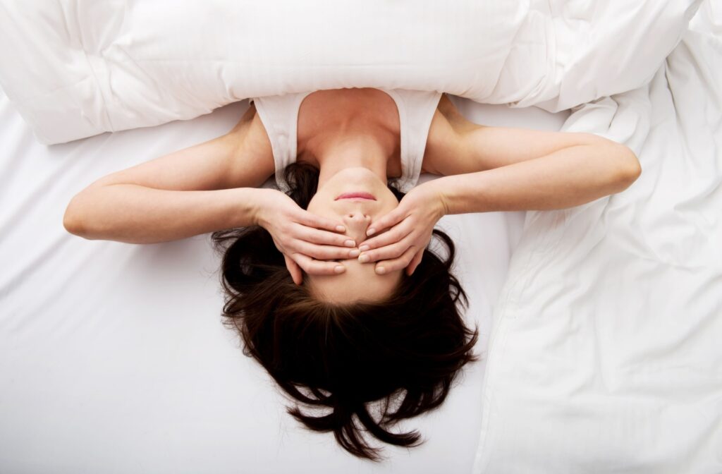 A young woman, laying on her bed, rubbing her dry eyes in the morning.